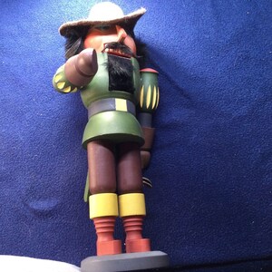 May include: A wooden nutcracker figurine with a white hat, brown beard, and green and brown clothing. The nutcracker is holding a gun in one hand and a sword in the other.