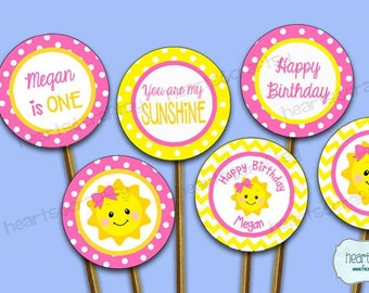 May include: A collection of six circular party decorations on wooden sticks. The decorations feature pink and yellow designs with text that reads "Megan is ONE", "You are my SUNSHINE", and "Happy Birthday". Some have a smiling sun graphic.