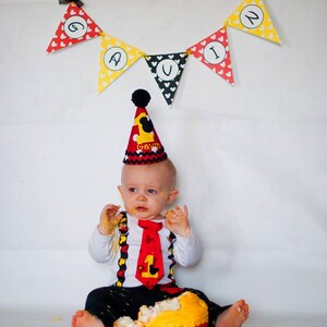 May include: A baby boy wearing a red and black Mickey Mouse themed birthday hat and outfit, with a red tie, is sitting on the floor with a yellow and red Mickey Mouse cupcake. A yellow and red banner with the name "Gavin" is hanging above the baby.