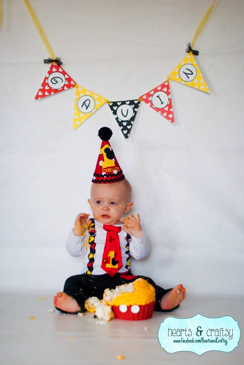 May include: A baby boy wearing a red and black Mickey Mouse themed birthday hat and outfit, with a red tie, is sitting on the floor with a yellow and red Mickey Mouse cupcake. A yellow and red banner with the name "Gavin" is hanging above the baby.