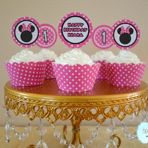 May include: Four pink and white polka dot cupcake wrappers with white frosting and a Minnie Mouse themed topper on each. The toppers say "Happy Birthday Kiara" and "1". The cupcakes are on a gold cake stand with crystal drops hanging from the bottom.