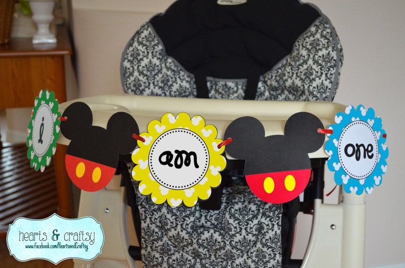 K&ouml;nnte beinhalten: Ein Mickey Mouse-Banner f&uuml;r eine erste Geburtstagsfeier. Das Banner hat abwechselnd Mickey Mouse-Ohren und Kreise mit den Worten "one" und "am" darauf. Das Banner ist an einem Hochstuhl befestigt.