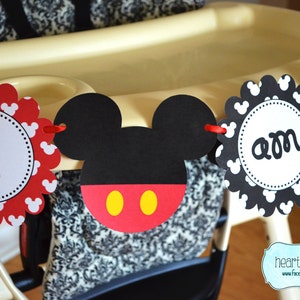 May include: A red, black and yellow Mickey Mouse themed banner with the words "I am one" on it. The banner is hanging from a high chair.