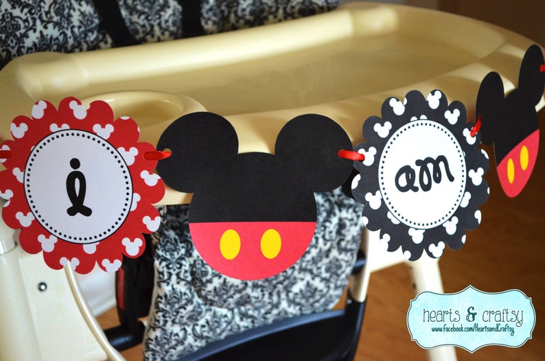 May include: A red, black and yellow Mickey Mouse themed banner with the words "I am one" on it. The banner is hanging from a high chair.