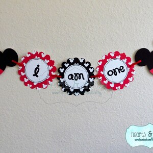 May include: A red, black and white Mickey Mouse themed banner with the words "I am one" on it. The banner is made of paper and has a scalloped edge.