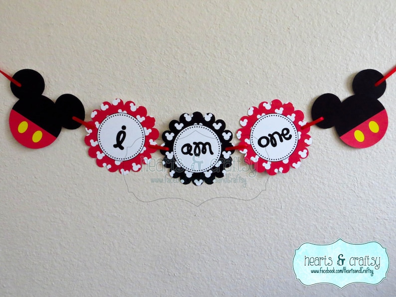 May include: A red, black and white Mickey Mouse themed banner with the words "I am one" on it. The banner is made of paper and has a scalloped edge.