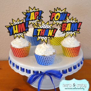 May include: A white cake stand with a blue ribbon and four cupcakes decorated with colorful superhero-themed toppers that say "Zap!", "Pow!", "Bam!" and "Yum!"  One cupcake has a topper that says "Ethan!"