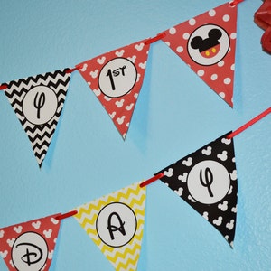 May include: A red, yellow, and black banner with Mickey Mouse ears and the words "Happy 1st Birthday" in white letters.