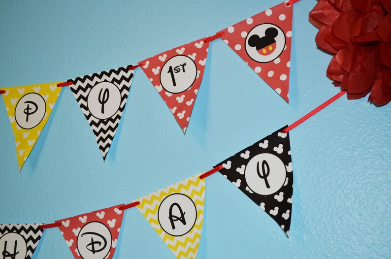 May include: A red, yellow, and black banner with Mickey Mouse ears and the words "Happy 1st Birthday" in white letters.