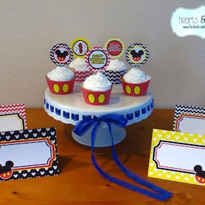 Può includere: Un set di topper per cupcake e segnaposto a tema Topolino. I topper presentano la testa di Topolino con orecchie rosse e nere e un cerchio giallo al centro. I segnaposto hanno un motivo a testa di Topolino bianco e nero con un bordo giallo e rosso. Il testo sui topper recita "Happy Birthday Cooper" e "Happy 1st Birthday".