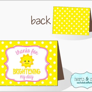 Pu&ograve; includere: Un biglietto giallo con pois bianchi e una cornice rosa. Il biglietto dice "grazie per aver illuminato la mia giornata" con un'immagine di sole sorridente.