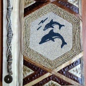 May include: A handmade photo album with a woven design featuring a brown, tan, and white color scheme. The album cover has a central hexagon with a silhouette of two dolphins swimming in a sandy beach scene. The album is bound with a brown leather strap and has a decorative metal button.