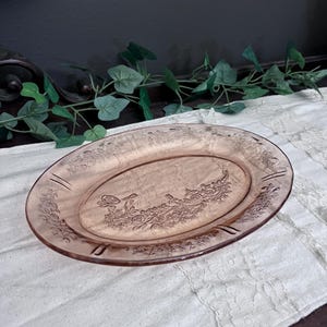 May include: An oval pink glass serving dish with an embossed floral design. The dish is displayed on a white lace tablecloth. The dish is a vintage item, likely from the mid-20th century.