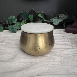 May include: A small, round, brass-colored container with a white, circular lid. The container has a slightly textured surface and sits on a white cloth. Decorative elements are visible in the background.