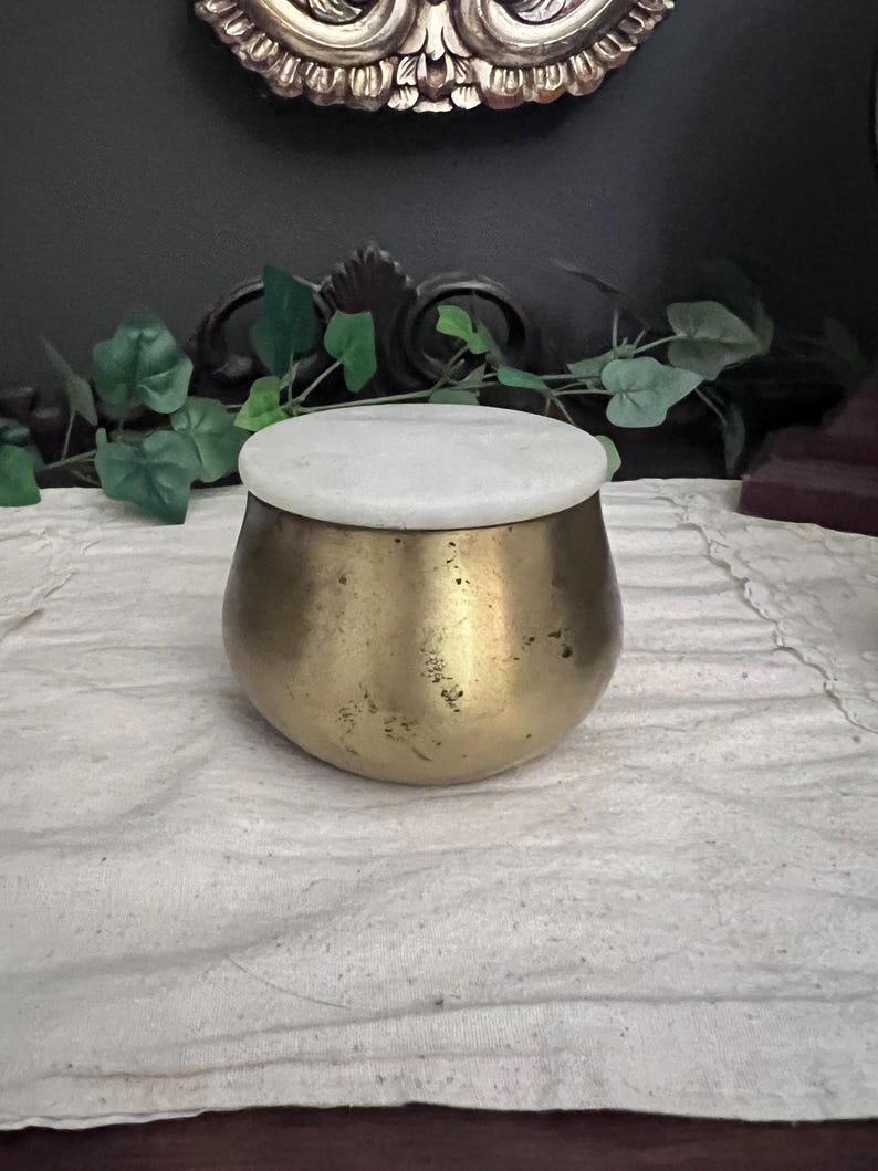 May include: A small, round, brass-colored container with a white, circular lid. The container has a slightly textured surface and sits on a white cloth. Decorative elements are visible in the background.