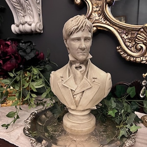 May include: A cream-colored bust of a man in a formal jacket and cravat, displayed on a silver-colored, ornate tray. The bust is set against a dark wall, with a decorative mirror and floral accents in the background.
