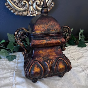 Op de afbeelding: Decoratieve doos in antieke stijl met deksel, in brons- en goudtinten. De doos heeft een verweerde afwerking, krulhandvatten en decoratieve accenten. Hij staat op kleine voetjes, met een bijpassende knop op het deksel.