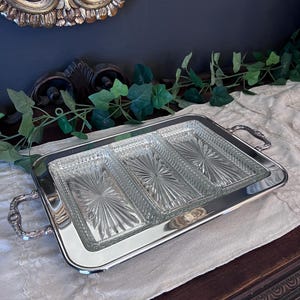 May include: A rectangular silver serving tray with three glass compartments. The glass compartments have a starburst pattern. The tray has ornate handles on each side. The tray is on a white surface.