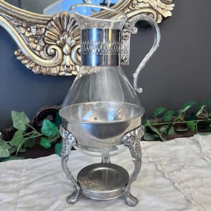 May include: A clear glass pitcher with a silver-toned metal collar featuring an embossed pattern and a curved handle. The pitcher rests in a silver-toned metal stand with three ornate, curved legs and a circular base designed for a warming candle.