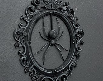 May include: A black spider in a black ornate frame with a decorative top. The frame is against a dark grey background.