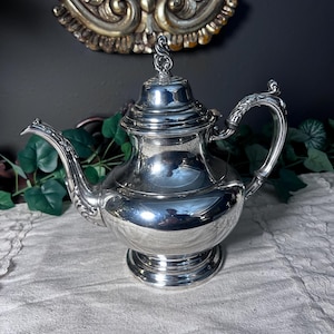 May include: A silver teapot with a curved spout and handle. The teapot has a decorative lid with a finial and sits on a round base. The teapot is highly polished and reflects the surrounding environment.