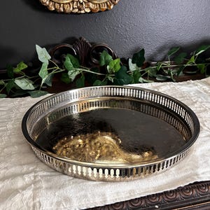 May include: A round silver-toned metal tray featuring a raised gallery rim with a repeating vertical cutout pattern. The interior base of the tray has a smooth, reflective surface that shows a distorted golden reflection of the surrounding environment.