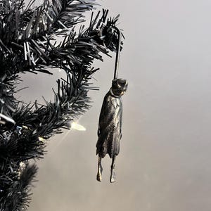 以下が含まれることがあります： 黒いクリスマスツリーに吊るされた、マントを着た人物の形をした暗い金属製のオーナメント。オーナメントは詳細なテクスチャで、金属製のフックで吊り下げられています。ツリーは暗く人工的な外観をしています。