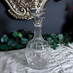 May include: A clear glass decanter with a stopper. The decanter has a round base with diamond-shaped cut glass patterns. The stopper is a flat disc shape. The background includes a gold-framed mirror and greenery.