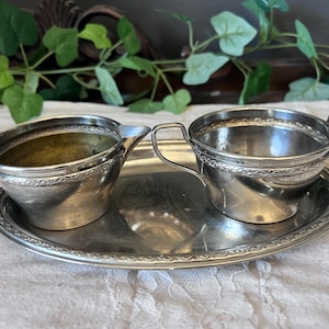 以下が含まれることがあります： 銀メッキのクリーマーと砂糖入れのセット、お揃いの楕円形のトレイ付き。縁とハンドル周りに装飾的なエンボス加工が施されています。クリーマーには注ぎ口があり、どちらのピースにも小さなハンドルが付いています。クラシックなサービングセット。