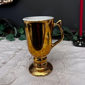 Puede incluir: Una taza dorada de estilo pedestal con interior blanco. La taza tiene una base redondeada, un tallo acanalado y un asa curva. La superficie refleja la luz, creando una apariencia brillante. La taza es probablemente de cerámica o metal.
