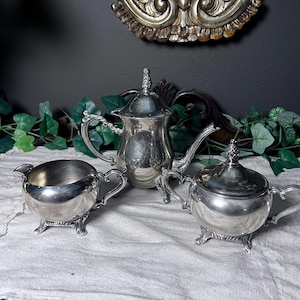 May include: A silver tea set, including a teapot, creamer, and covered sugar bowl, is displayed on a white cloth. The set has ornate detailing and sits on a dark wooden surface. A decorative silver piece is in the background.
