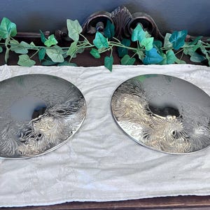 May include: Two circular silver-toned metal plates featuring a polished center and an intricate, embossed floral scrollwork pattern around the outer rim. The plates have a reflective, metallic finish and a smooth, rounded edge design.
