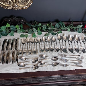 May include: A collection of silver-colored flatware, including spoons, knives, and forks, arranged on a white tablecloth. The silverware is displayed on a dark wooden surface, with decorative elements in the background. A red candle is visible.