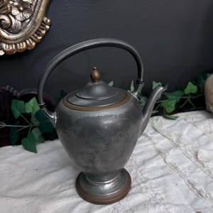 May include: Antique metal teapot with a curved handle and a small spout. The teapot has a dark gray finish with copper-colored accents around the base and lid. The teapot is sitting on a white, textured surface.