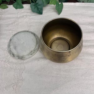 May include: A small, antique brass bowl with a matching lid. The bowl has a rounded shape and a slightly tarnished finish, giving it a vintage appearance. The lid is made of a light-colored stone. The bowl sits on a white lace cloth.