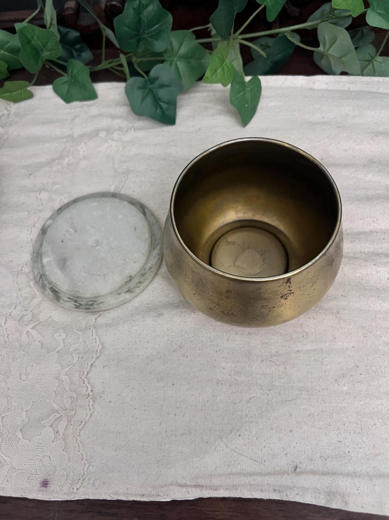 May include: A small, antique brass bowl with a matching lid. The bowl has a rounded shape and a slightly tarnished finish, giving it a vintage appearance. The lid is made of a light-colored stone. The bowl sits on a white lace cloth.