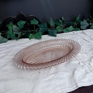 Op de afbeelding: Roze ovale glazen schaal met een getextureerde, ruitvormige rand en een geribbelde binnenkant. De schaal wordt gepresenteerd op een wit, getextureerd oppervlak. De schaal is waarschijnlijk vintage en geschikt om te serveren of tentoon te stellen.