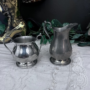 May include: Two antique silver-colored serving pieces. A small, round creamer with two handles and a pedestal base sits next to a taller, pitcher-shaped creamer with a handle and a spout. Both are on a white lace surface.
