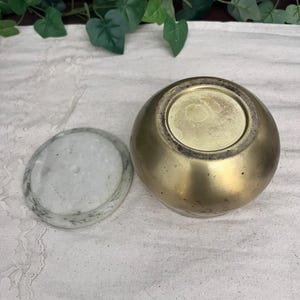 May include: A small, round, brass-colored container with a matching lid. The lid is a flat, circular piece of white and gray stone. The container is sitting on a white cloth.