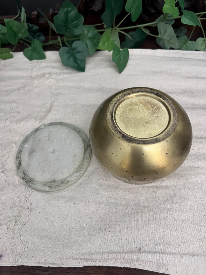 May include: A small, round, brass-colored container with a matching lid. The lid is a flat, circular piece of white and gray stone. The container is sitting on a white cloth.