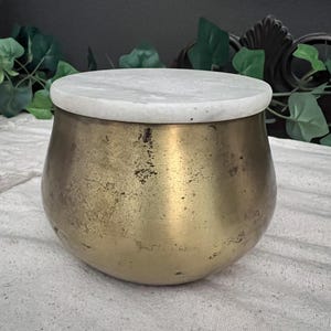 May include: A round, antique brass container with a white marble lid. The container has a bulbous shape and a tarnished, aged appearance. The lid is circular and sits flush on top of the container. The background includes green foliage.
