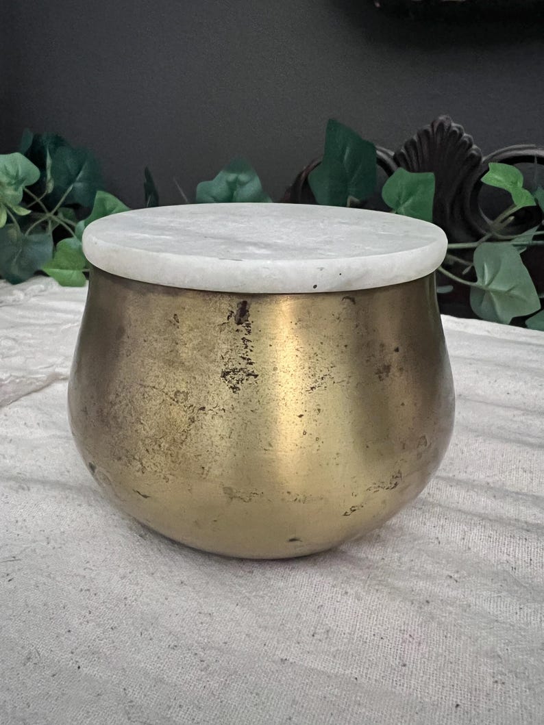 May include: A round, antique brass container with a white marble lid. The container has a bulbous shape and a tarnished, aged appearance. The lid is circular and sits flush on top of the container. The background includes green foliage.