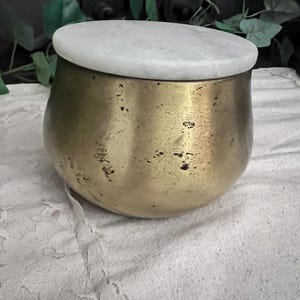 May include: A small, round, brass-colored container with a white marble lid. The container has a slightly textured surface and a rounded shape. The lid is flat and circular, contrasting with the metallic base.