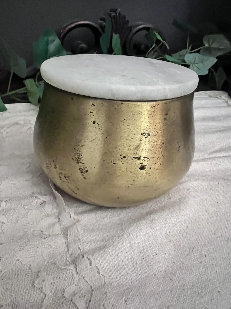 May include: A small, round, brass-colored container with a white marble lid. The container has a slightly textured surface and a rounded shape. The lid is flat and circular, contrasting with the metallic base.