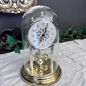 May include: A gold-tone anniversary clock with a glass dome, featuring a white circular face with black Arabic numerals and a central black floral pattern. The clock has a rotating pendulum with four gold-tone spheres and a brass-colored base.
