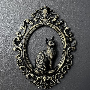 May include: Ornate, oval-shaped wall decor featuring a seated cat sculpture. The cat is dark, with a detailed texture, set within a gold-toned frame with intricate scrollwork and a crown-like top. The background is a textured gray wall.