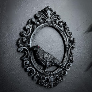 May include: A black raven sculpture with red eyes is centered within an ornate, oval-shaped frame. The frame and raven are dark gray, with intricate detailing. The background is a solid, dark gray.