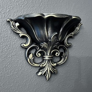 May include: A wall-mounted shelf featuring a scalloped, bowl-shaped top and an ornate, symmetrical base with scrollwork and fleur-de-lis detailing. The piece is finished in a dark black color with metallic gold highlights on the raised edges.
