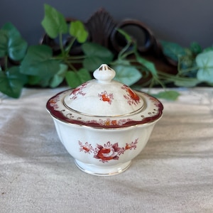 May include: A white ceramic sugar bowl with a lid, featuring a floral pattern in shades of red and maroon. The bowl has a scalloped edge with a maroon border and gold trim. The lid has a small finial.