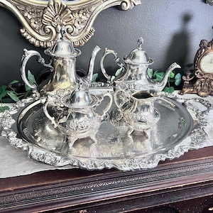 Pode incluir: Um conjunto de chá em metal prateado composto por um bule grande, um bule pequeno, um açucareiro com tampa e uma leiteira, todos com detalhes florais em relevo e pegas ornamentadas, dispostos sobre um tabuleiro oval a combinar.